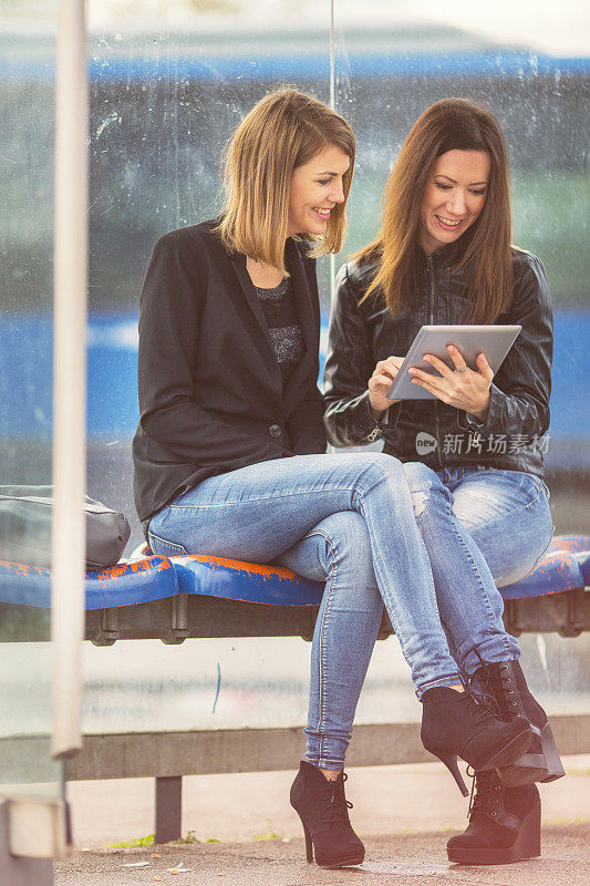 Two friends using digital tablet on bus station